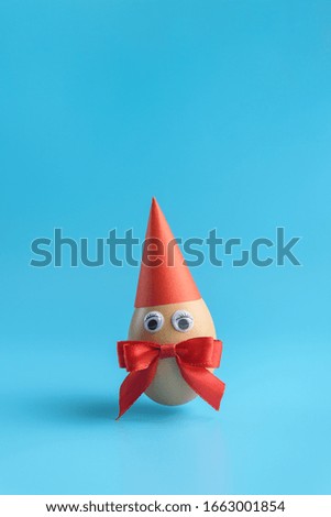 One brown egg with eyes, a red ribbon bowin and a red festive cap on a blue background. Minimal Happy Easter concept decoration. Copy space for text mockup. Close up photography.