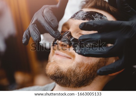 Barber applies black charcoal mask to man face to clean pore skin and remove acne from nose, spa salon. Royalty-Free Stock Photo #1663005133