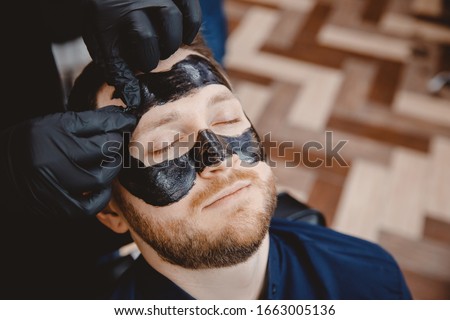 Acne removal with cosmetic product, cleaning pores of men skin, barber applying black charcoal mask to client man in spa salon. Royalty-Free Stock Photo #1663005136