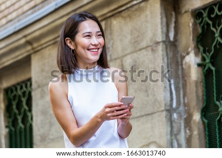 Portrait of a young, attractive and beautiful Asian woman smiling as she texting on her smartphone int he city. She is looking away as she talks animatedly and happily on her mobile device.