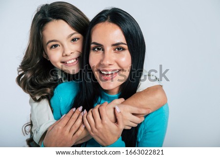 Love and affection. Close-up photo of a young cheerful mother and her little cute daughter, who is hugging her from the back, while they both are looking in the camera and smiling.