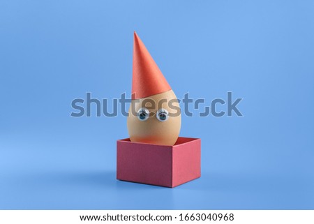 One brown egg with eyes in a red festive cap in a square red box on a blue background. Minimal Easter concept decoration. Copy space for text mockup. Close up photography.