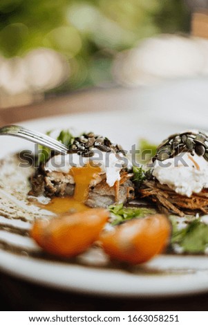 Food photo of shrimp with poached eggs, black seeds, sauce, parsley and red tomato on white plate on blurred green background