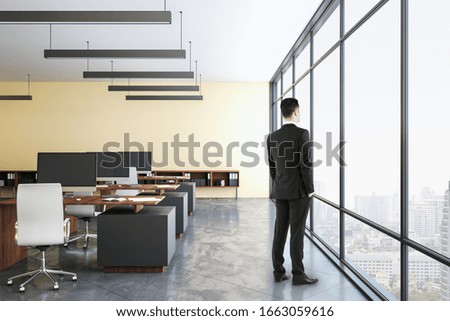 Businessman standing in office interior with computer monitor and panoramic city view.