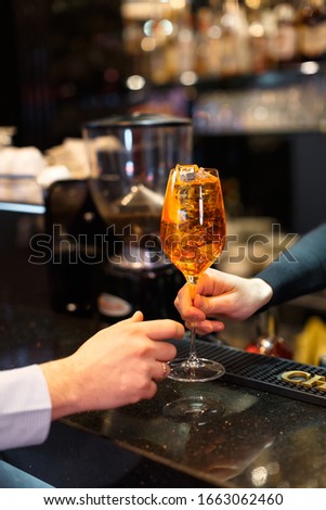 Person hands an orange aperol cocktail to another person. Classical italian aperitif. Alcoholic beverage with lots of ice. Vertical close up photo. Selective focus on glass, blurred background.
