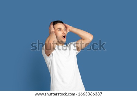 Astonished, shocked. Caucasian young man's portrait isolated on blue studio background. Beautiful male model in casual style, pastel colors. Concept of human emotions, facial expression, sales, ad.