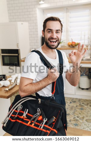 Image of young caucasian plumber man in uniform holding bag with equipment and gesturing okay sign while working in apartment