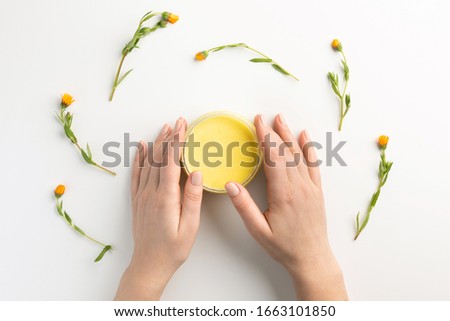 Organic cosmetic concept. Female hands, flowers of calendula and jar of skin care cream, isolated on white background. Flat lay, top view, copy space.