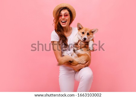 Cute brunette woman in white t shirt and jeans holding and embracing corgi puppy dog on plane pink background. Love to the animals, pets concept. cheerful woman holding Welsh Corgi dog. Happy together