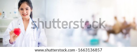 Beautiful smiley doctor hold red heart shape in hand at hospital with blur people background, healthcare and medical concept.