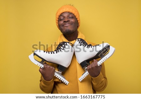 African young guy in hat and sweater and scarf, holds skates in his hands smiling at camera on yellow background. Winter hobby for health concept.