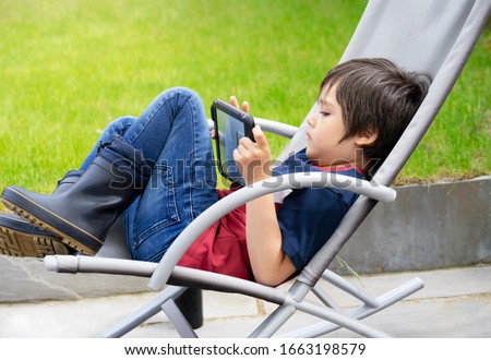 Portrait kid with playing game on tablet, A boy having fun watching cartoons on digital tablet, Child siting on rocking chair relaxing on weekend in the garden on spring or Summer, Home schooling
