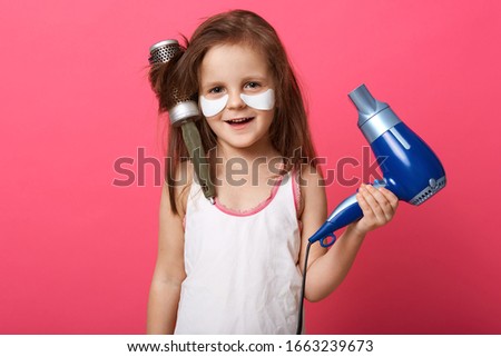 Studio shot of charming cute female kid posing isolated over rose studio background with blue hairdryer in hands, little girl with combin her hair, having tangled hairbrush on head. Children concept.
