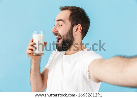 Portrait of a young cheerful excited bearded man wearing t-shirt standing isolated over blue background, showing glass of milk while taking a selfie