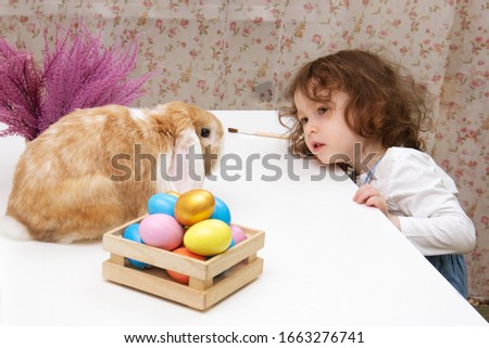 easter bunny and a little baby girl are looking at each other. The hare is sitting on the table, the girl is smiling. colorful eggs on purple flowers background provence. Copy space for text.