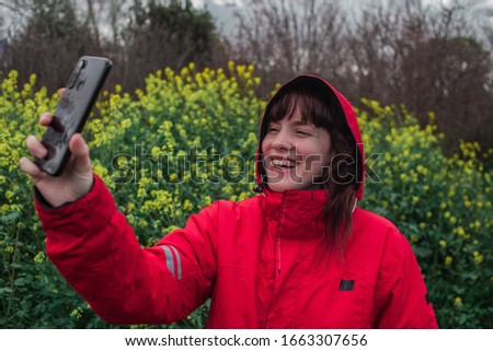 Young woman with red pea coat taking a picture with mobile phone while smiling in the field