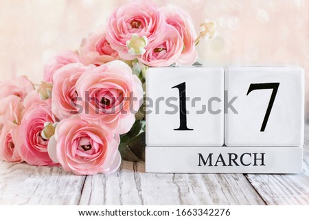 White wood calendar blocks with the date March 17th and pink ranunculus flowers over a wooden table. Selective focus with blurred background. St Patrick Day.