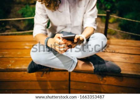 Young woman sitting relaxed on wooden bench with her legs crossed holding mobile device in her hands looking at screen. Girl using smart phone summer night at green park.