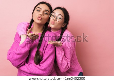 Image of two happy teenage girls with braids in casual clothes smiling and blowing air kiss to you isolated over pink background