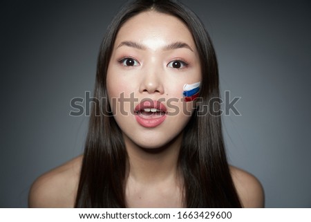  Portrait of a young beautiful Asian girl with a Russian flag painted on her cheek, looking directly at the camera, surprised. On a gray background, a place to copy. The concept of patriotism, footbal