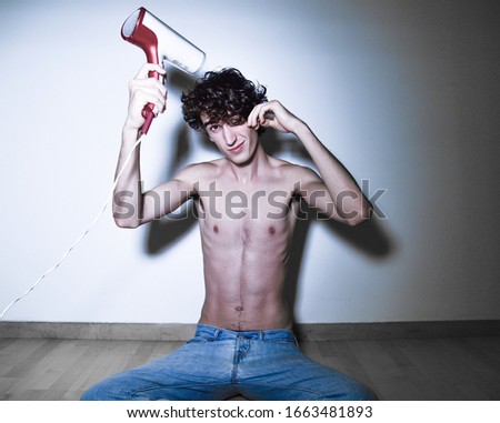 portrait of expressive mediterranean Italian dark haired handsome male model boy with angel face and fit sportive shaped body playing with hair blower , sitting on wooden floor with white background