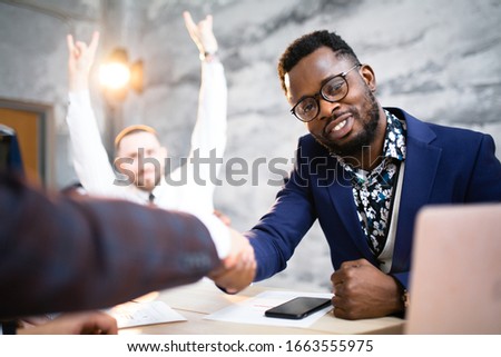 An African American in a blue suit shakes hands with a business partner as a sign of cooperation between the two firms. Joyful worker in the background