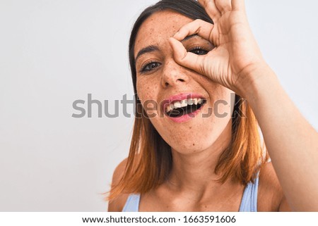 Beautiful redhead woman standing over isolated background with happy face smiling doing ok sign with hand on eye looking through fingers