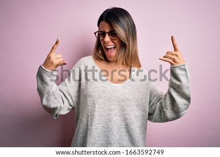Young beautiful brunette woman wearing casual sweater and glasses over pink background shouting with crazy expression doing rock symbol with hands up. Music star. Heavy concept.
