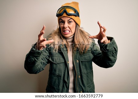 Young brunette skier woman wearing snow clothes and ski goggles over white background Shouting frustrated with rage, hands trying to strangle, yelling mad