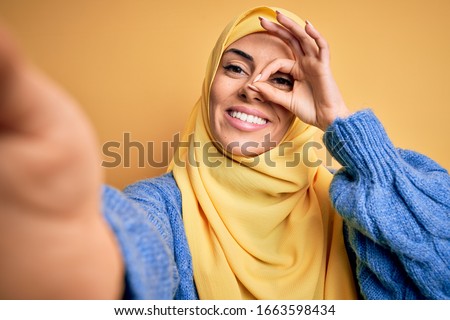 Young beautiful brunette arab woman wearing islamic hijab make selfie by camera with happy face smiling doing ok sign with hand on eye looking through fingers