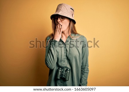 Beautiful blonde explorer woman with blue eyes wearing hat and glasses using binoculars bored yawning tired covering mouth with hand. Restless and sleepiness.