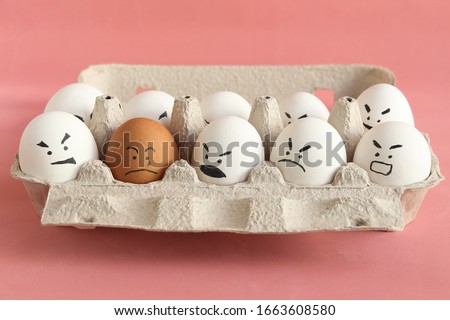 Group of white organic chicken eggs with angry faces and one brown chicken egg with crying face in carton box made from recycled paper. Red background. Theme of racism and intolerance in society. Royalty-Free Stock Photo #1663608580