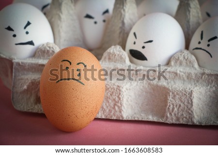 A brown egg with a painted crying face stands next to a recycled paper box containing white eggs with angry faces. Red background. Theme of racism and intolerance in society. Royalty-Free Stock Photo #1663608583