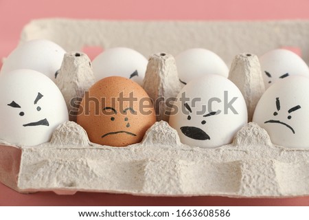 Group of white organic chicken eggs with angry faces and one brown chicken egg with crying face in carton box made from recycled paper. Red background. Theme of racism and intolerance in society. Royalty-Free Stock Photo #1663608586