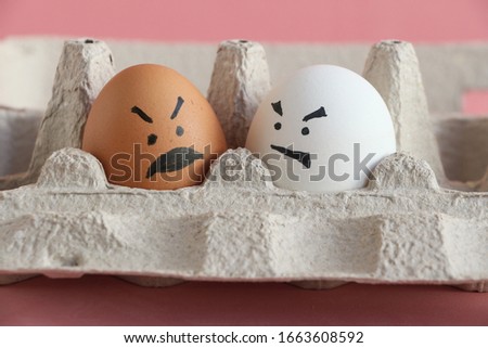 White organic chicken egg next to brown chicken egg with in carton box made from recycled paper. Angry faces painted on both eggs. Theme of racism and intolerance in society. Royalty-Free Stock Photo #1663608592