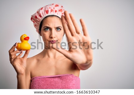 Young brunette woman with blue eyes wearing bath towel and shower cap holding duck toy with open hand doing stop sign with serious and confident expression, defense gesture