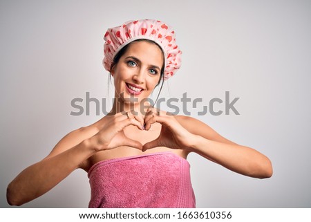 Young brunette woman with blue eyes wearing bath towel and shower cap smiling in love showing heart symbol and shape with hands. Romantic concept.