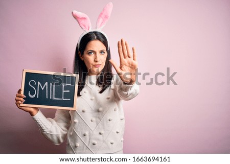Young caucasian woman wearing easter rabbit ears and holding blackboard with smile word with open hand doing stop sign with serious and confident expression, defense gesture