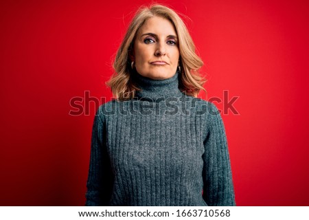 Middle age beautiful blonde woman wearing casual turtleneck sweater over red background with serious expression on face. Simple and natural looking at the camera.