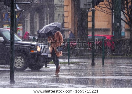 A woman in a brown coat under an umbrella crosses a busy street in heavy rain. Heavy precipitation in the city. Flooding of city streets. Royalty-Free Stock Photo #1663715221