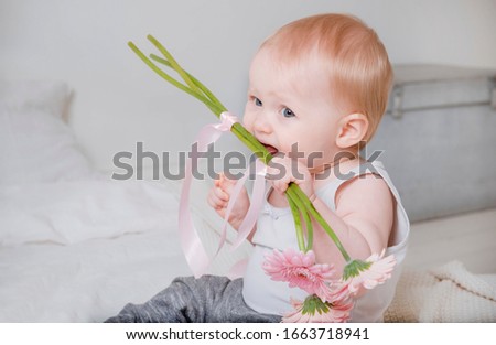1 little blonde girl sitting on the bed in a light top and gray pants and biting flower stalks, pink gerberas, close up
