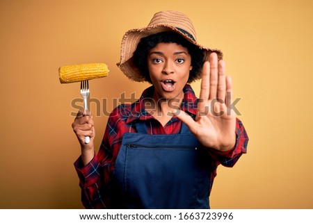 Young African American afro farmer woman with curly hair wearing apron holding cob corn with open hand doing stop sign with serious and confident expression, defense gesture