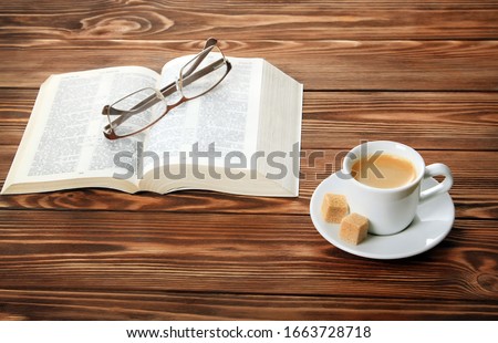 An open book on the table, glasses, a cup of cappuccino coffee with sugar cubes, blue background.