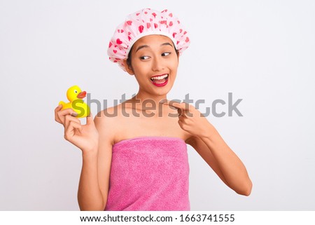 Chinese woman wearing shower towel and cap holding duck toy over isolated white background very happy pointing with hand and finger