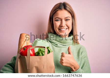 Young beautiful blonde girl holding fresh groceries paper bag over pink isolated background happy with big smile doing ok sign, thumb up with fingers, excellent sign