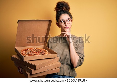 Young beautiful woman with curly hair and piercing holding delivery boxes with Italian pizza serious face thinking about question, very confused idea