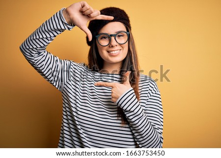 Young beautiful brunette woman wearing french beret and glasses over yellow background smiling making frame with hands and fingers with happy face. Creativity and photography concept.