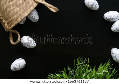 Top view of easter arrangement. White eggs with painted silver details, scattered from jute bag on black background and green plant. Copy space in the middle. Traditional holiday season.