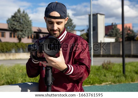 Caucasian young man with reverse cap and red jacket works outdoor with his camera on a gimbal stabiliser. Video operator focusing with his video camera. Cinema lens on DLSR camera. Medium angle shot