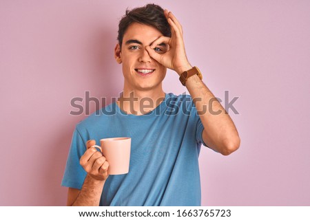 Teenager boy drinking a cup of coffee over isolated pink background with happy face smiling doing ok sign with hand on eye looking through fingers